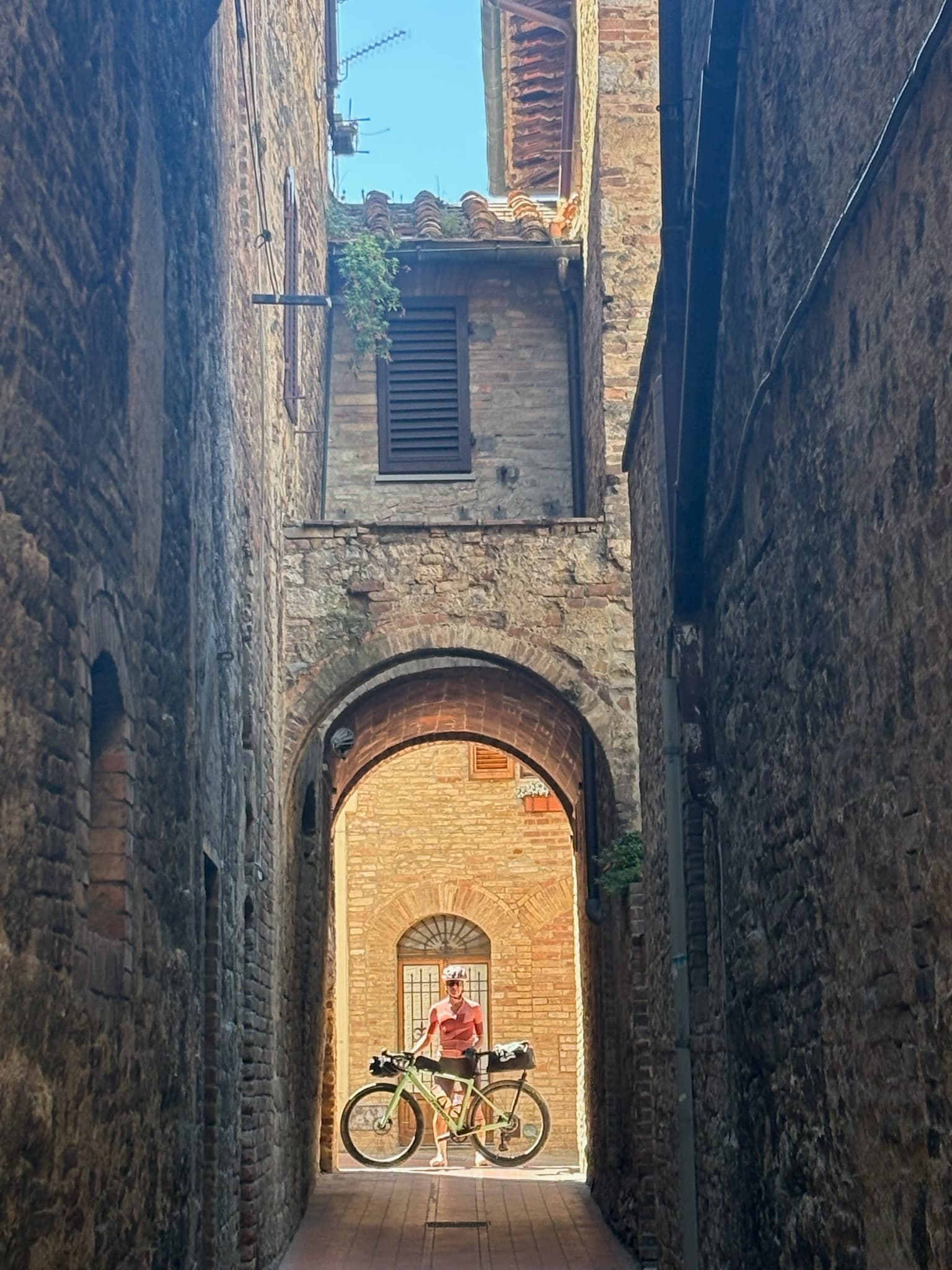 A cyclist in a helmet and sunglasses standing with a loaded gravel bike under a sunlit brick archway, framed by a narrow, shadowed stone alley in an Italian hill town.