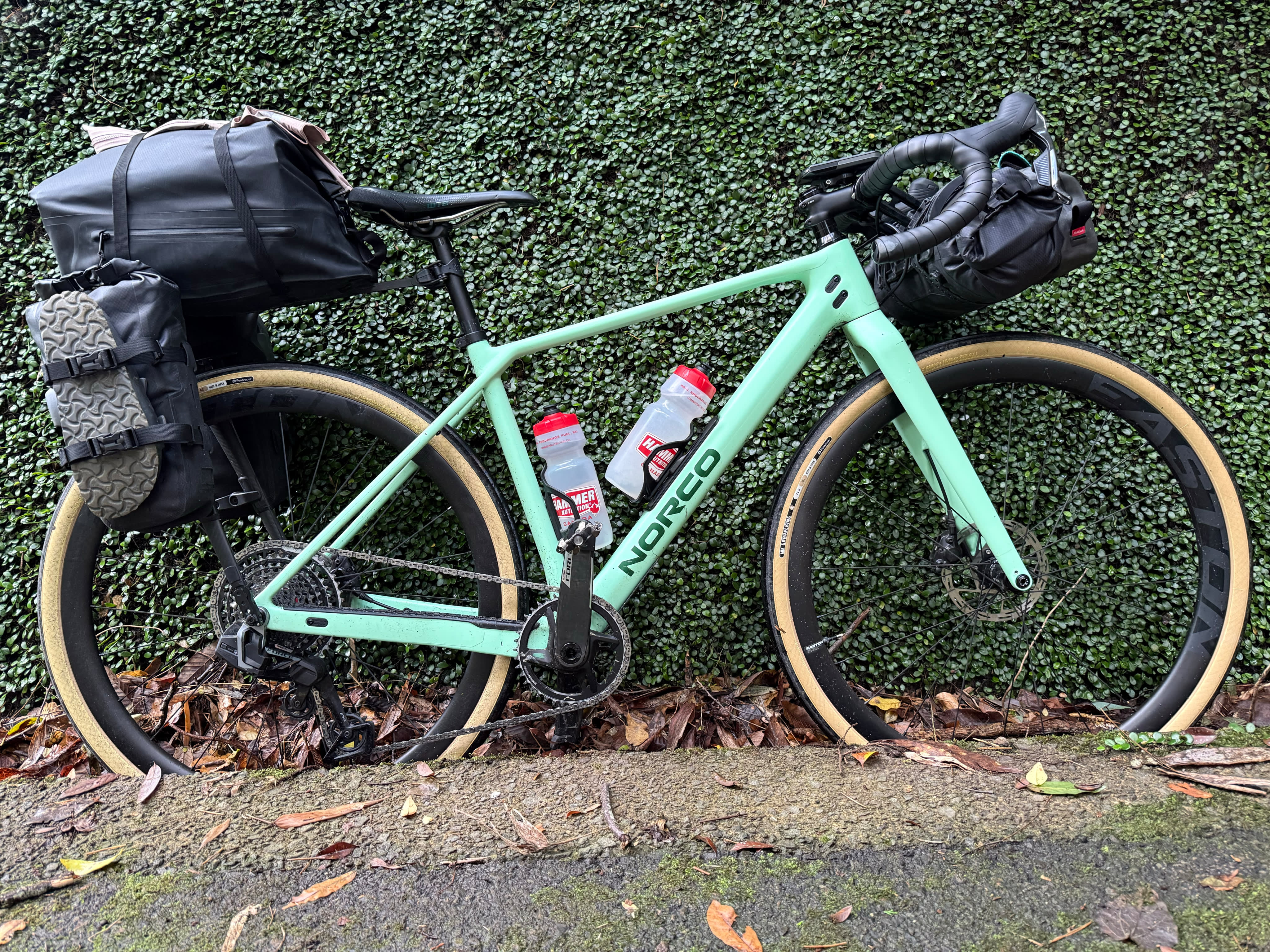 A mint green Norco gravel bike leaning against a hedge, loaded with black bikepacking bags on the handlebars, frame, and seat post, two water bottles mounted on the frame.