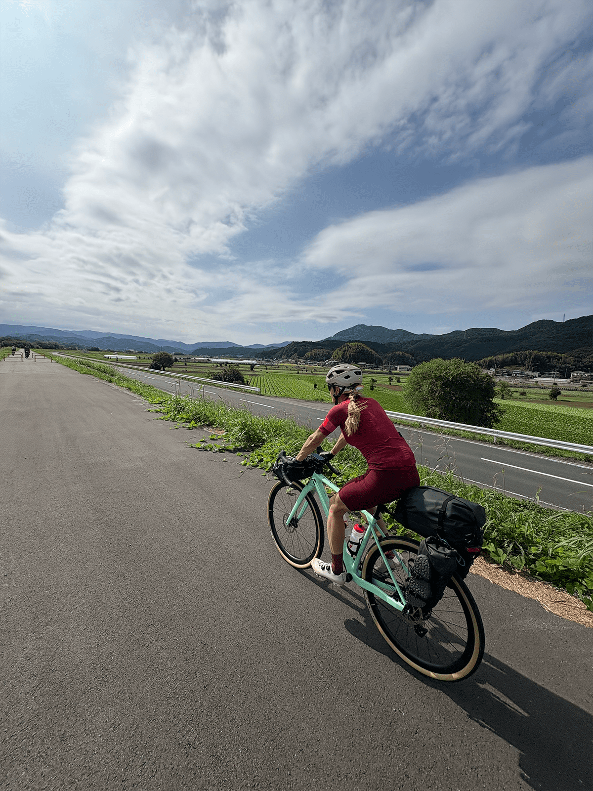 René Unser riding a mint green loaded touring bike along a paved road beside rice paddies and fields, mountains in the background, Japan.