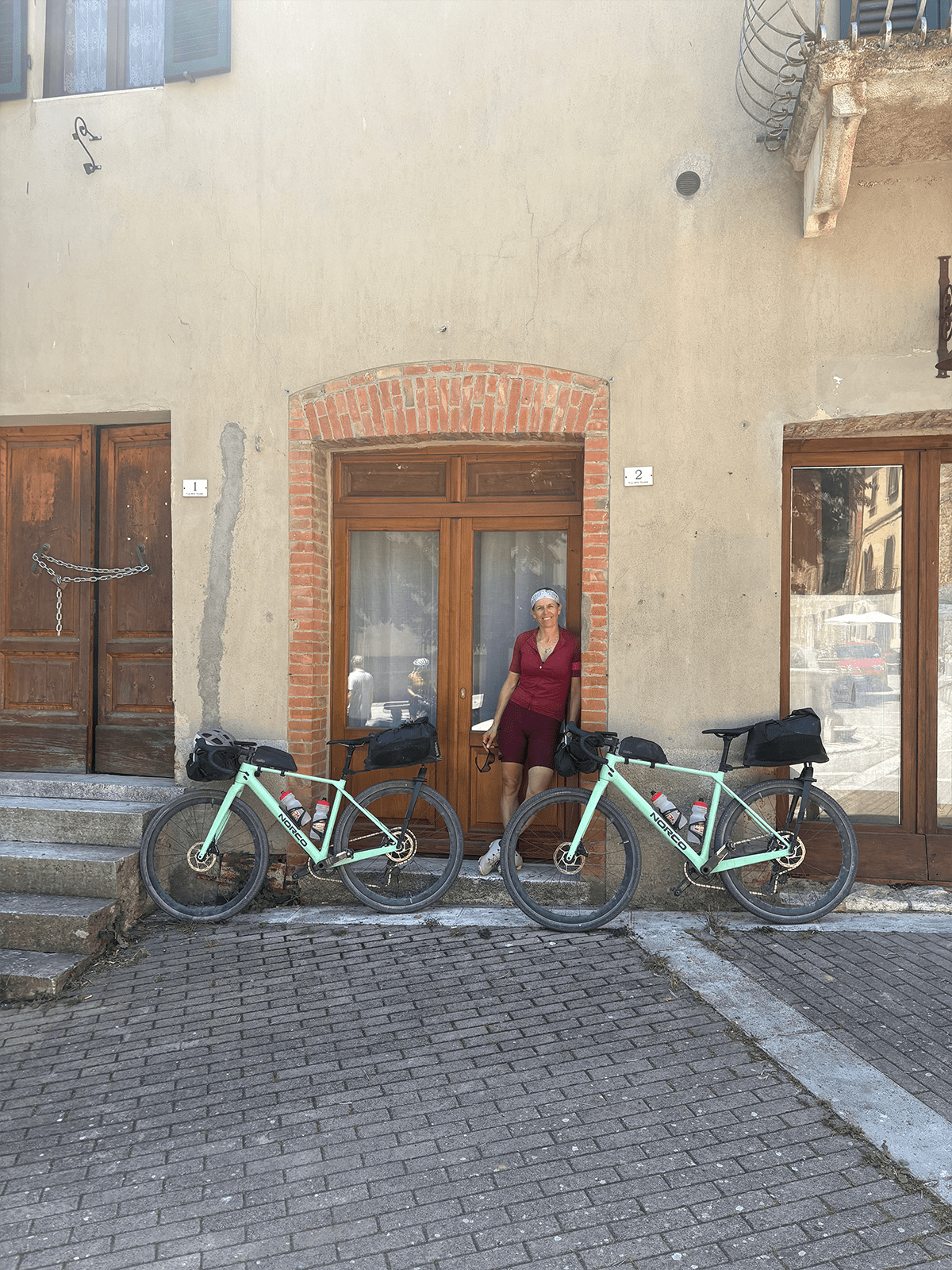 René Unser standing between two mint green Norco touring bikes with bikepacking bags, parked outside an old stone building with wooden doors in Tuscany, Italy.
