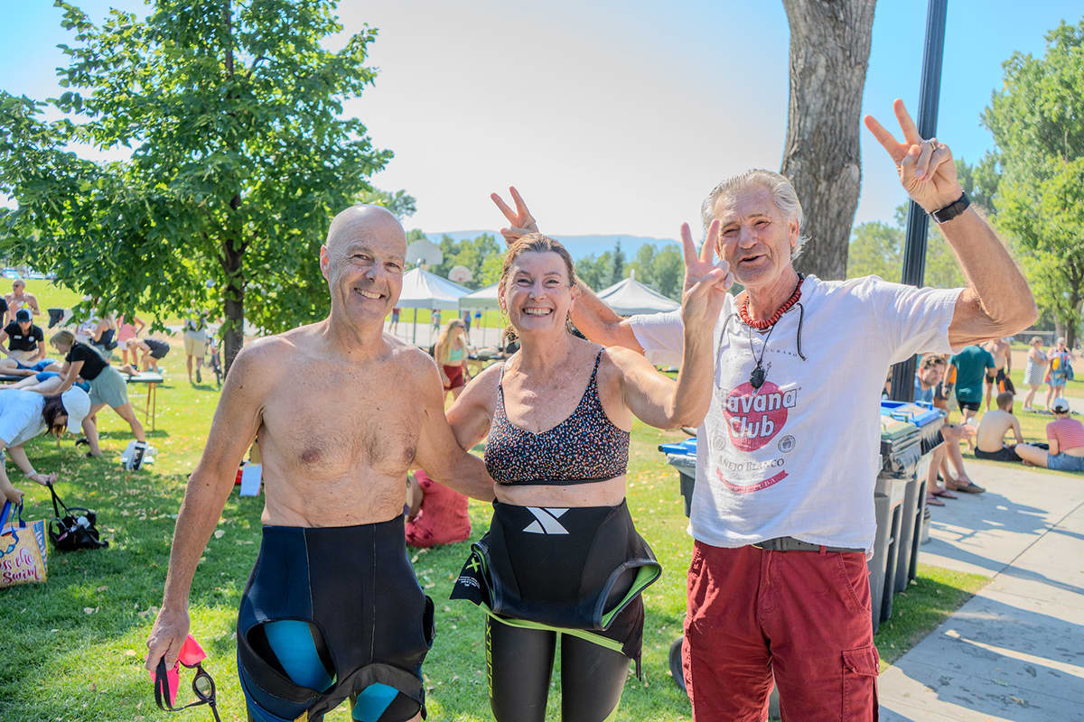 Three adults in partially removed wetsuits pose together laughing at the finish area, with recovery tents and a park setting behind them.