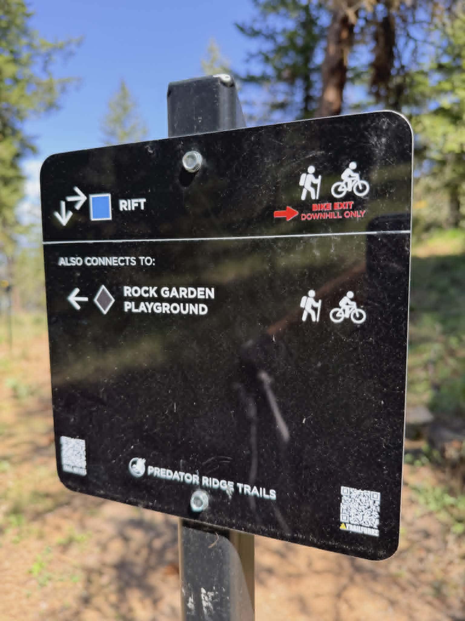 Black Predator Ridge trail sign showing the Rift trail (blue rating, bike exit downhill only) with an arrow pointing left to Rock Garden Playground, Trailforks QR code at bottom right Photo: Cody Cosens | Predator Ridge Trails