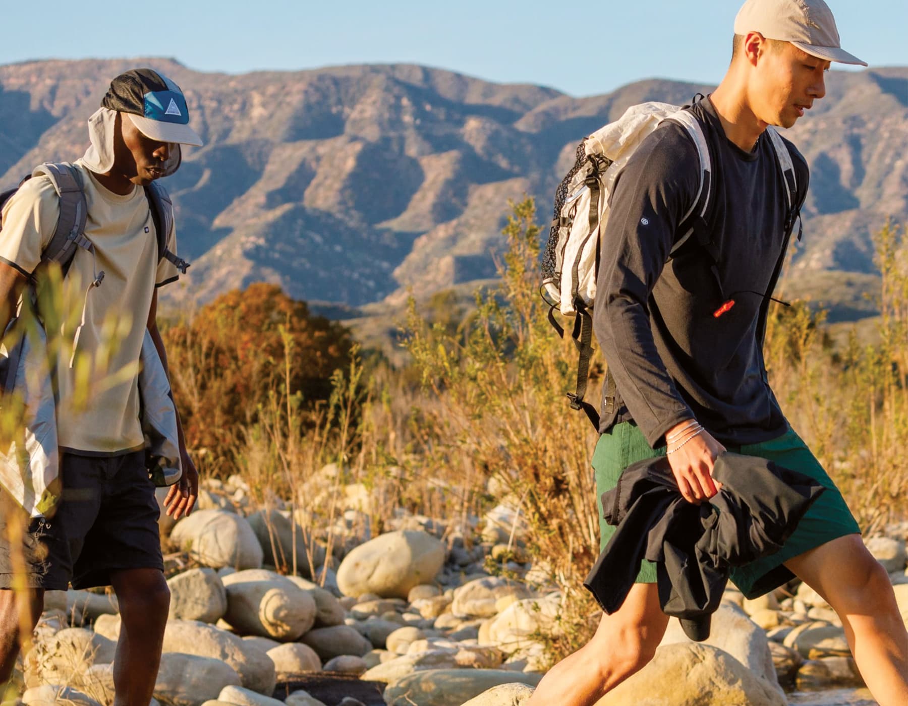 Two male hikers walking on a trail with small rocks and tall bushes with a mountain backdrop. Both are wearing shorts, backpacks and hats on the sunny day outside. 