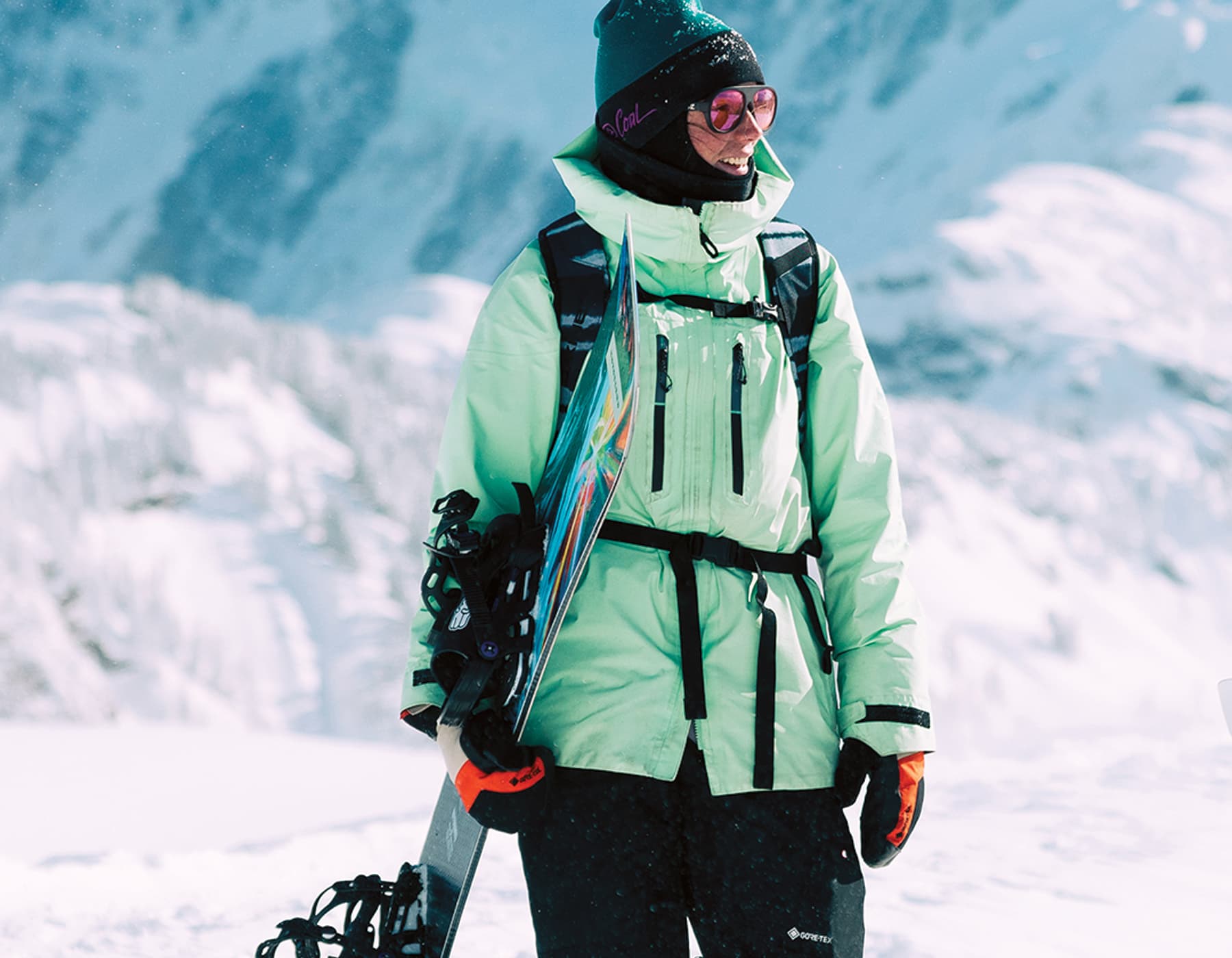 Female snowboarder standing in the snow high uptop a sunny mountain holding a snowboard. She is wearing a green 686 jacket, mittens, winter hat and glasses.