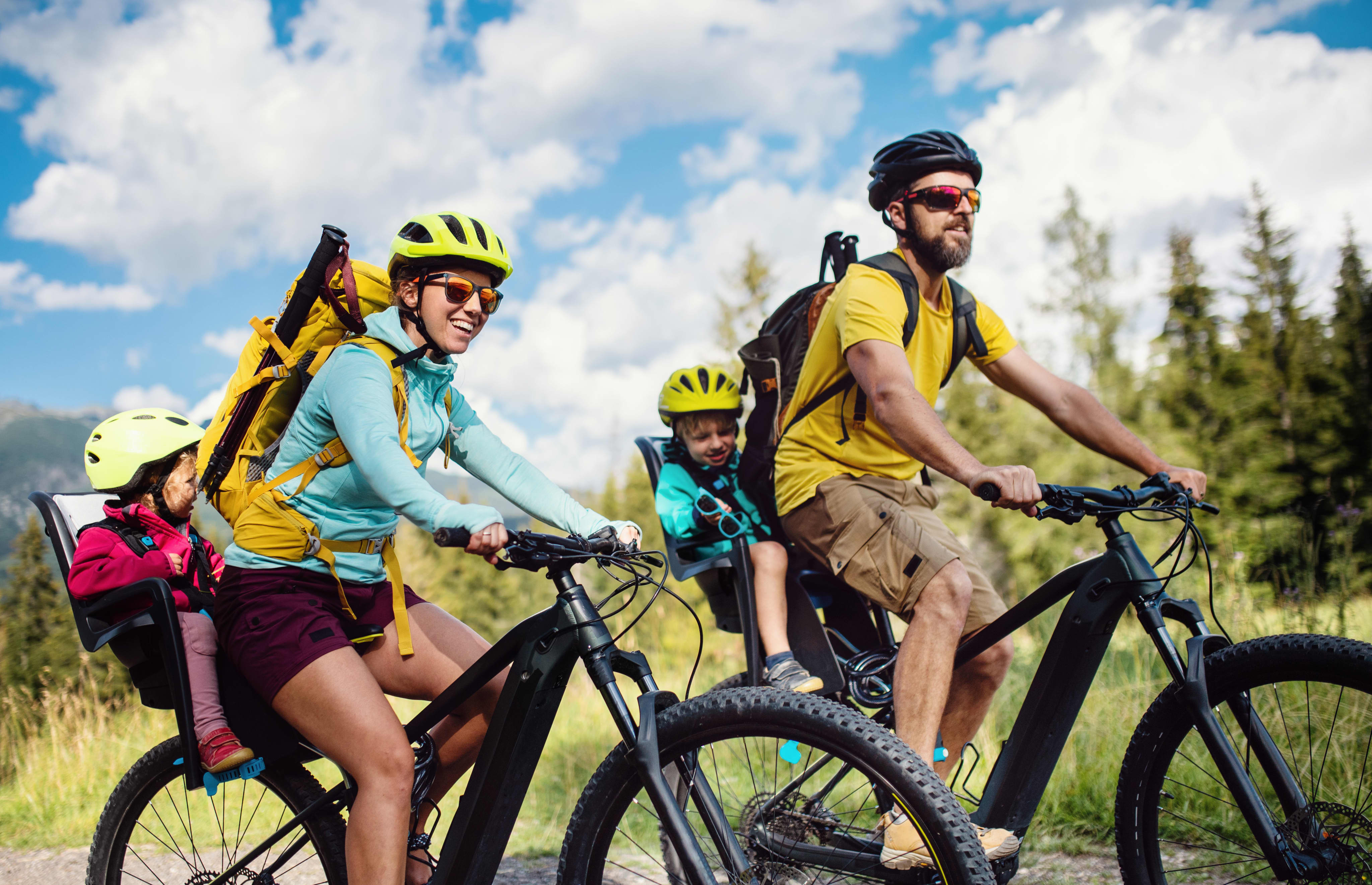 Two adults and two young children ride mountain bikes together on a sunny trail surrounded by trees and mountains. Both adults wear helmets and sunglasses; the children ride in rear-mounted seats.