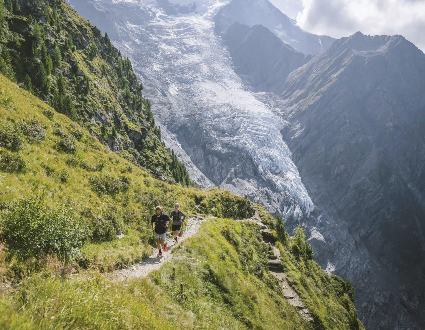 Two runners along a steep mountain running path. A lush green mountainside with a snowy, rocky mountain in the background. Both runners are wearing Altra running shoes and hydration vests.