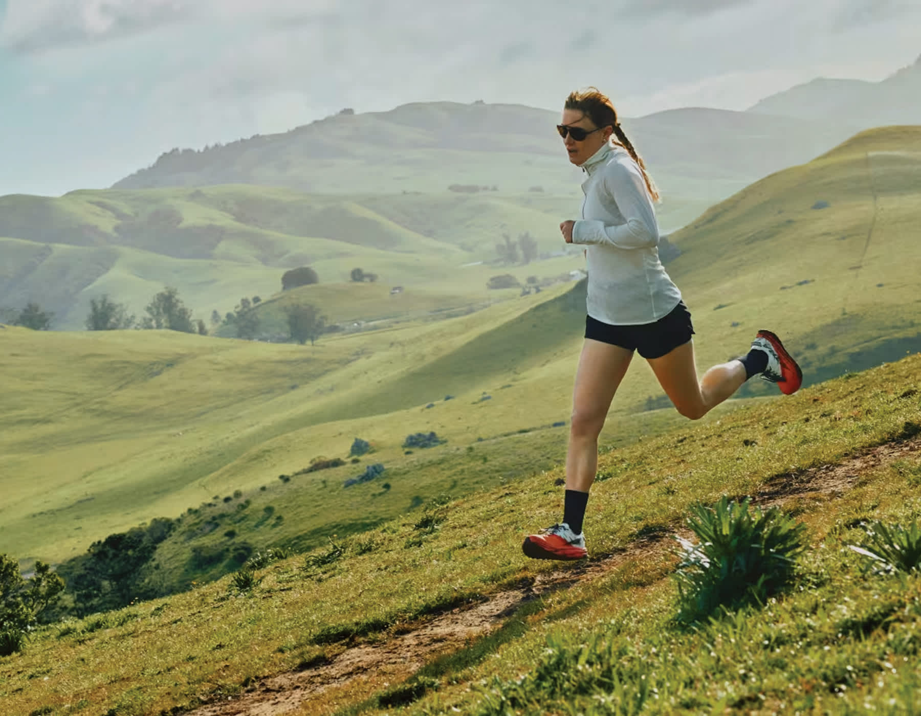 Woman Runnign along a hillside wearing Altra running shoes, women's running apparel and sunglasess. The hillside is lush and green with clouds in a blue sky.