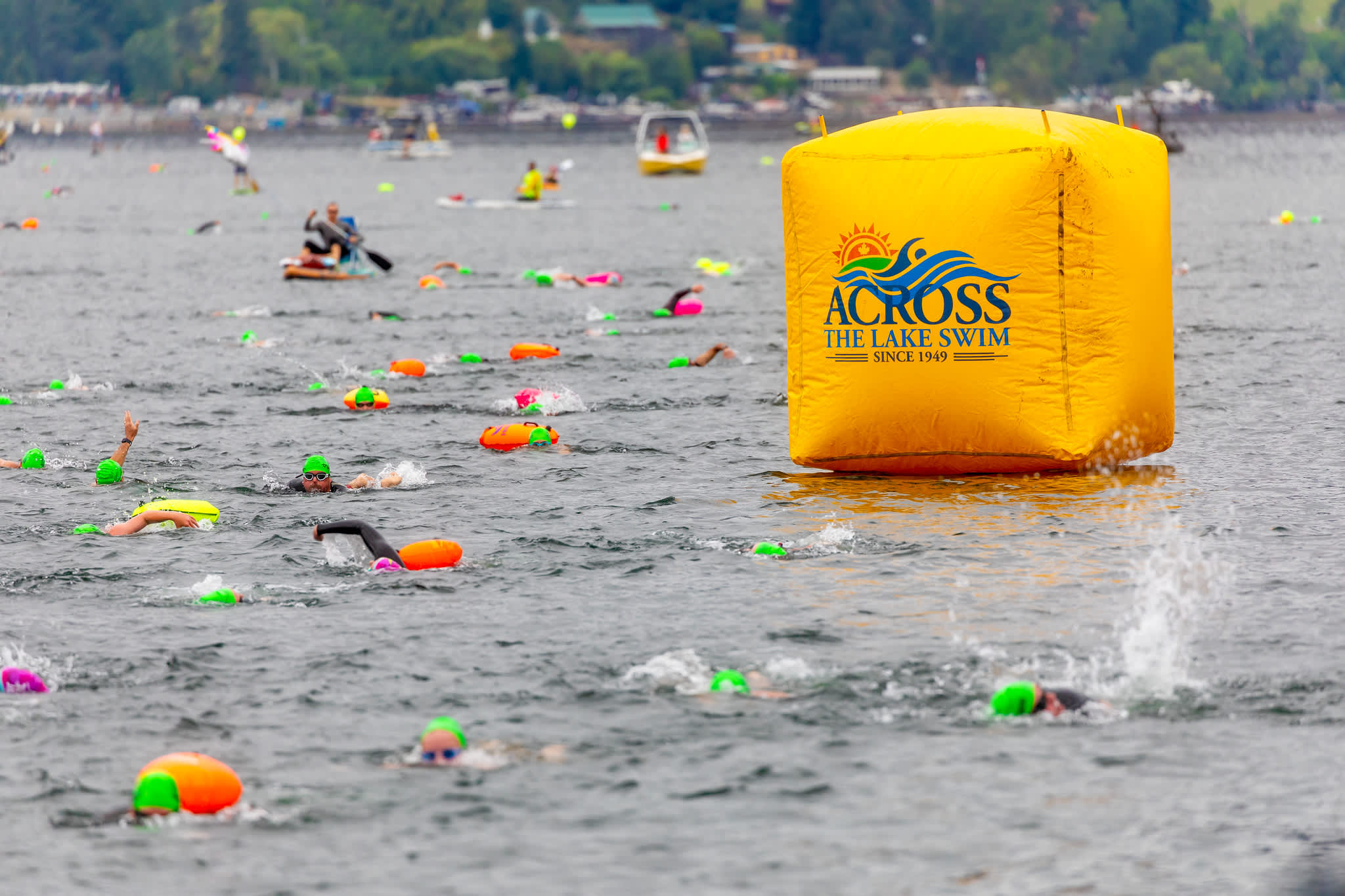 Open water swimmers in bright caps and tow floats pass a large yellow Across the Lake Swim buoy, with support boats and paddleboarders nearby.
