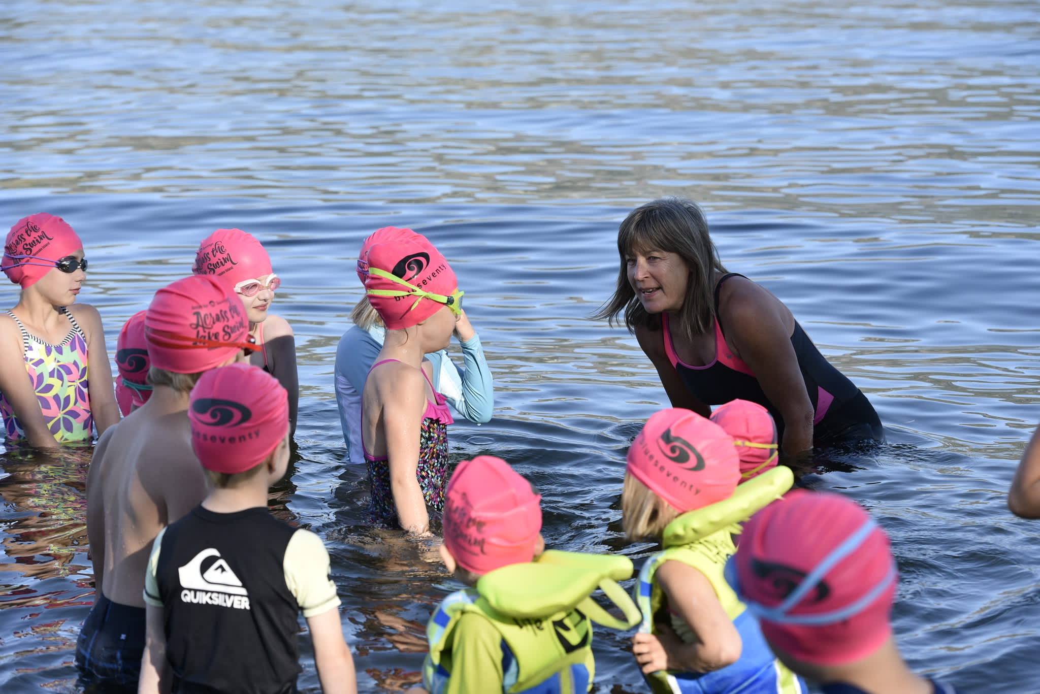  A coach crouches in shallow water talking to children wearing pink Across the Lake Swim swim caps and tow floats.