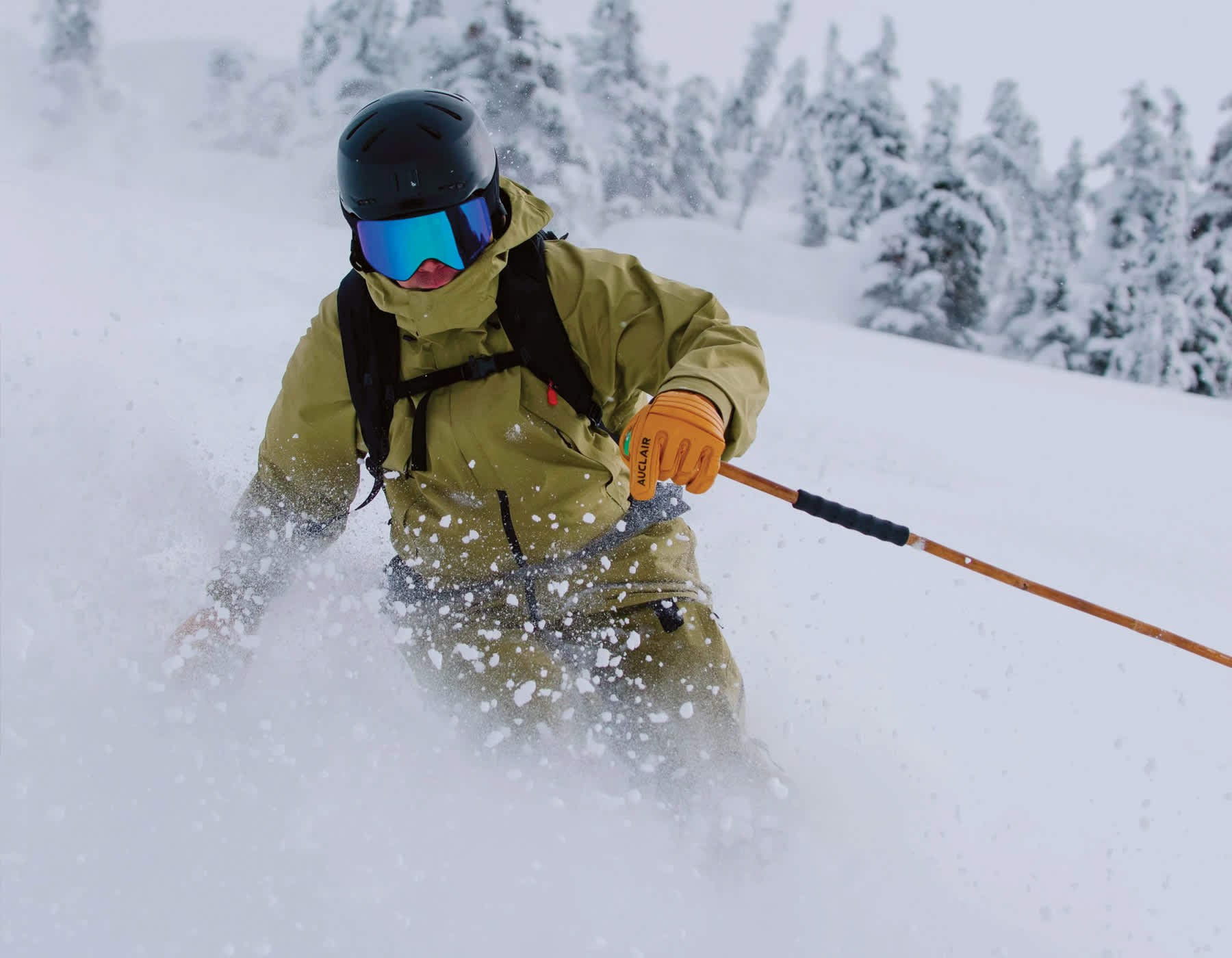 An Auclair athlete sking through powder snow and it flies up in front of him with speed. He is wearing a green jacket, camel colored Auclair gloves while holding ski poles mid turn.