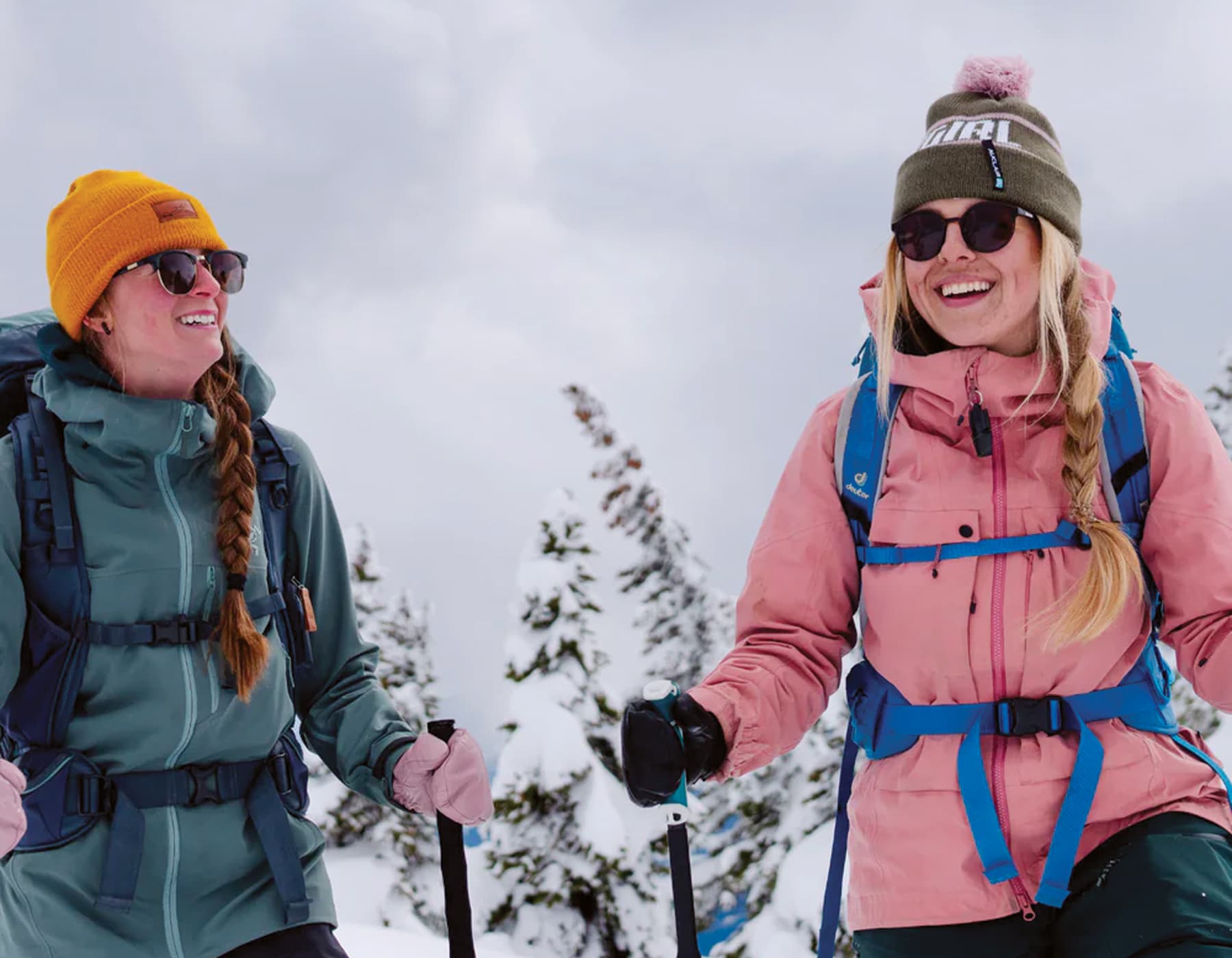 Two females snowshoeing, laughing and having a good time outside in the snow. They are both wearing backpacks with the female on the left wearing a green jacket and orange beanie looking at the women on the right wearing a pink jacket and green beanie. Both women are holding poles and wearing dark sunglasses. 