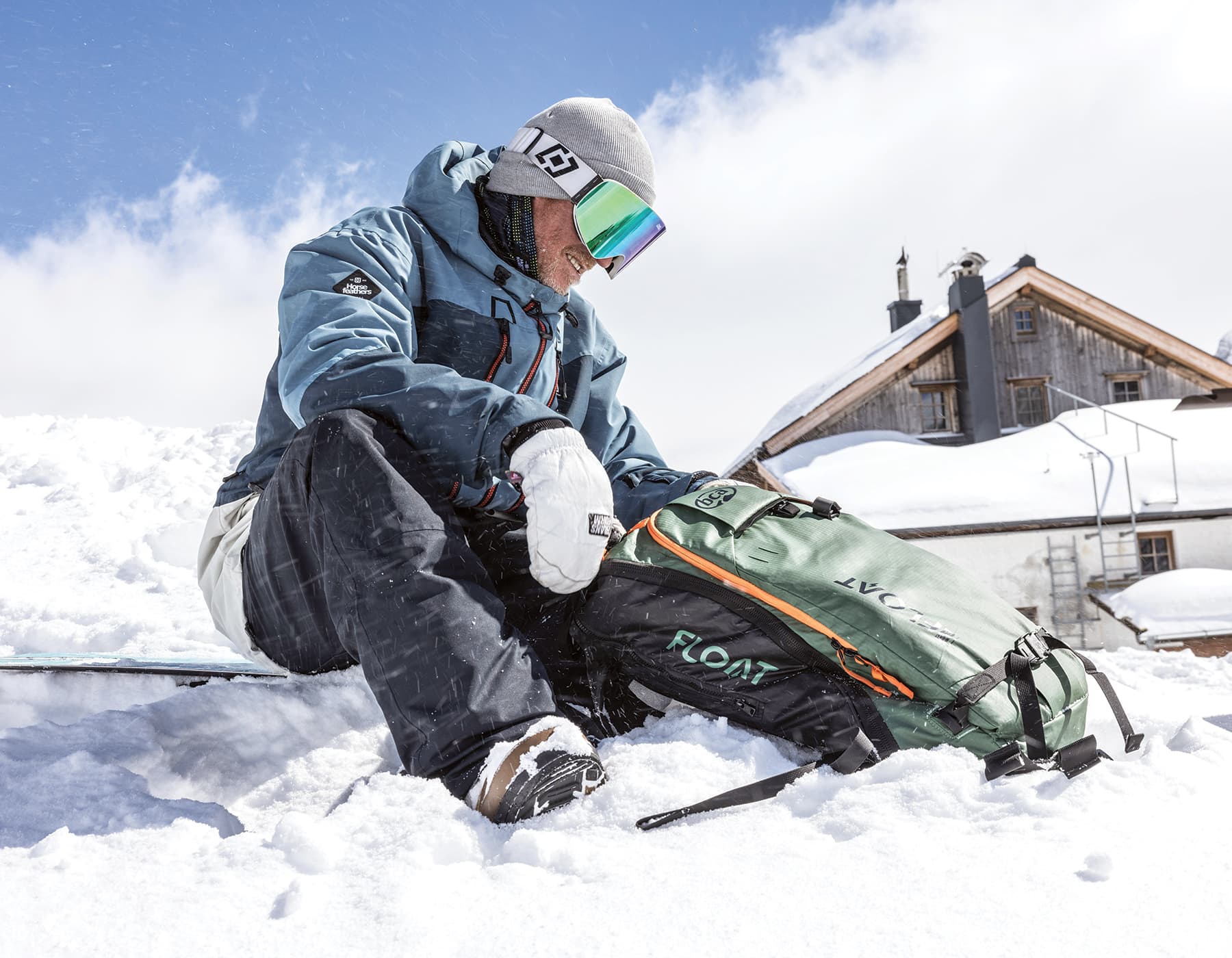 Snowboarder sitting outside of a snowy capped backcountry cabin. He is wearing full ski gear with goggles and gloves looking down at a Backcountry Access Float backpack. 