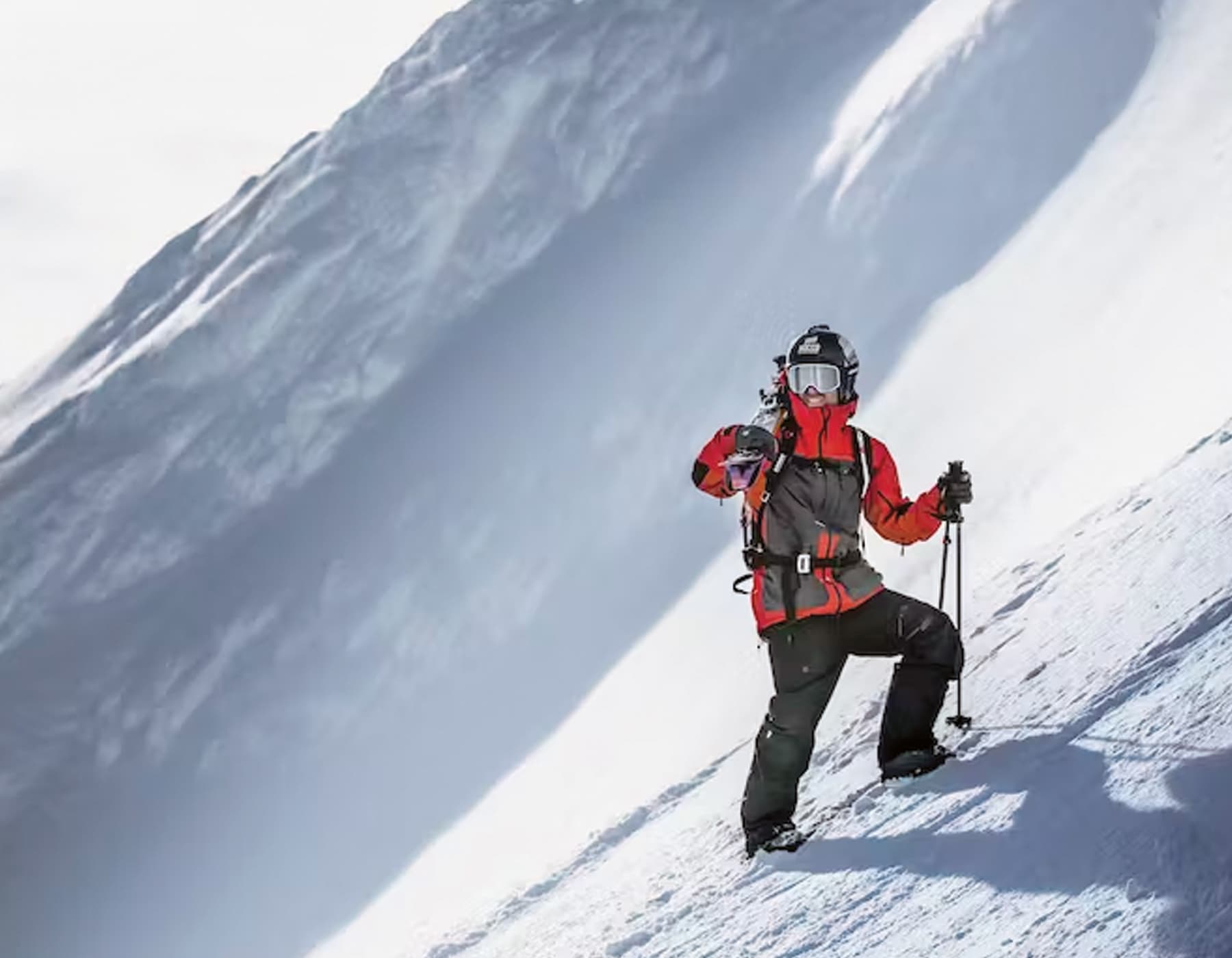 Hestra Athlete Chad Sayers standing on a steep mountain side in full ski gear, helmet and goggles carrying his skis and poles.