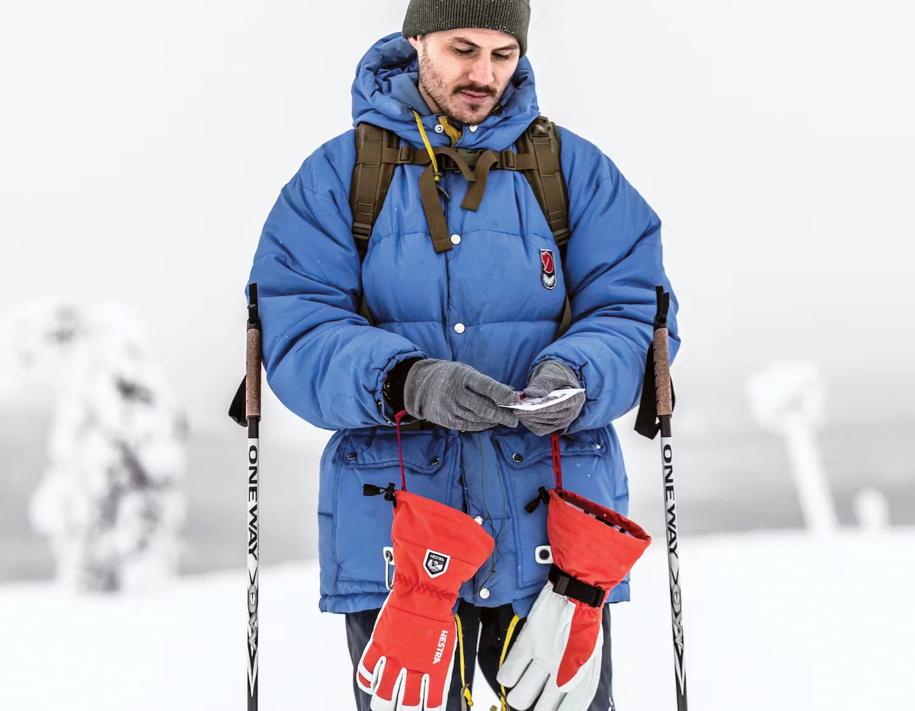A skier in a blue jackets standing in the snow with 2 poles and a backpack. He is looking at a piece of paper while a pair of red Hestra gloves hangs from his wrists. 