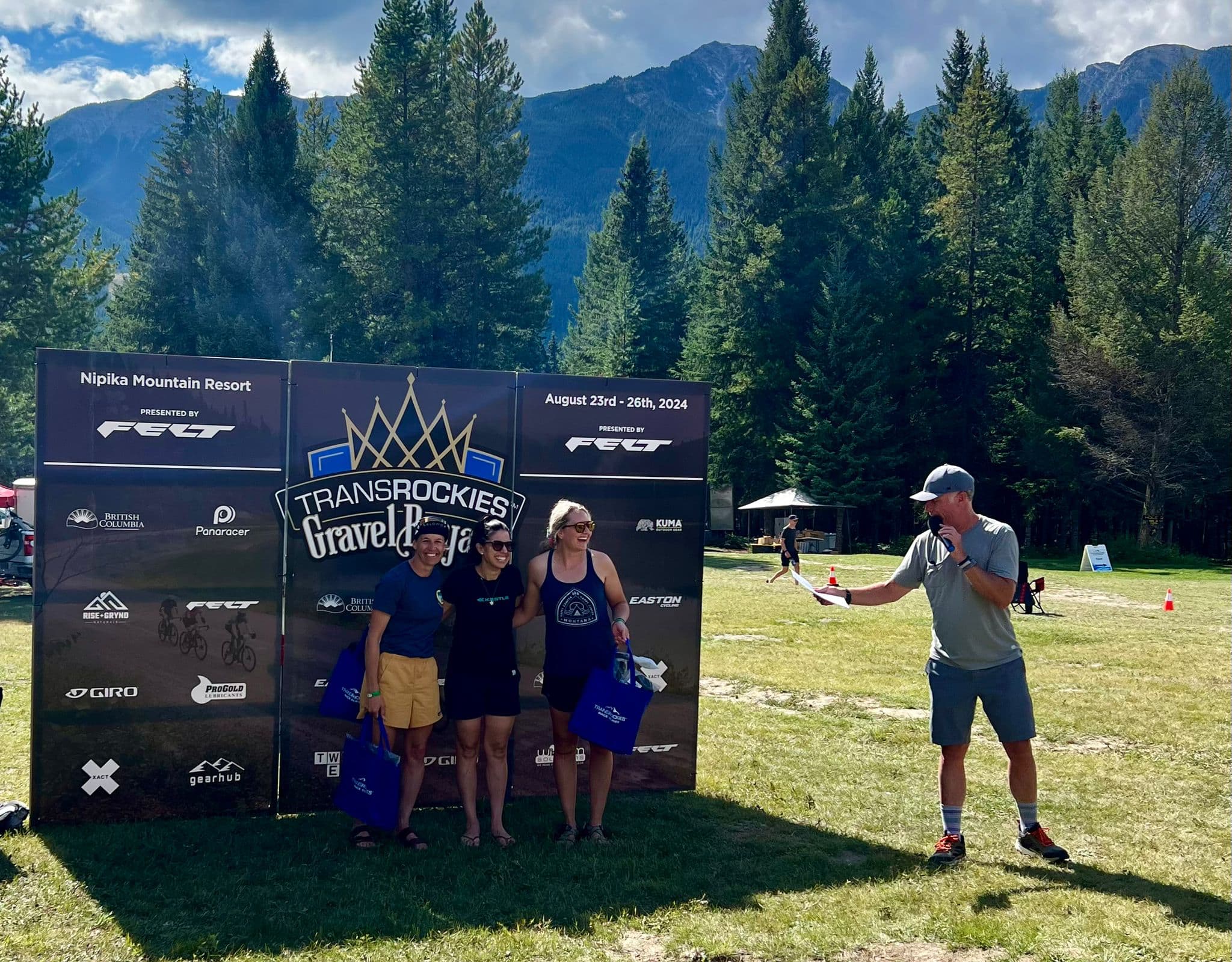 Three women posing beside a TransRockies Gravel Royale event sign at Nipika Mountain Resort, holding event tote bags, with an announcer speaking into a microphone and mountains in the background.