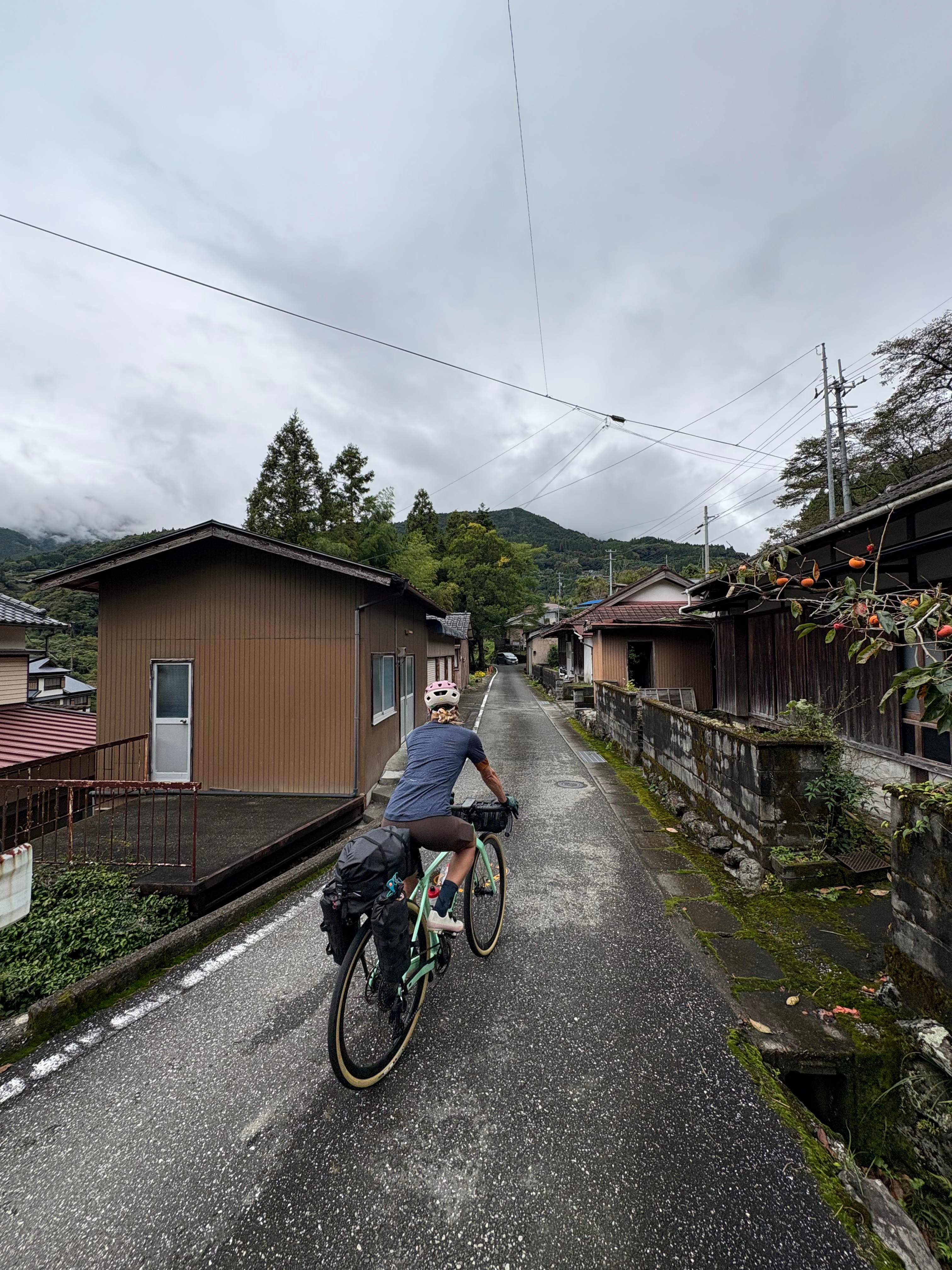 René Unser riding a mint green loaded touring bike away from the camera down a narrow paved road between traditional houses, forested mountains visible ahead under an overcast sky, Japan.