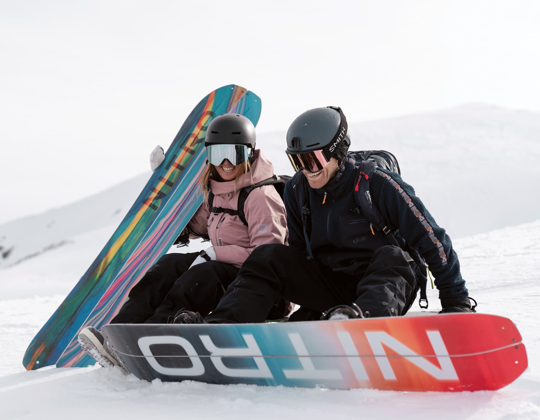 Female snowboarder wearing a pink jacket, helmet and goggles sitting in the snow on the side of a run, smiling and holding a Nitro snowboard. Beside her is a male snowboarder wearing a black jacket, helmet and goggles with his feet strapped into a Nitro snowboard, also sitting and smiling. 