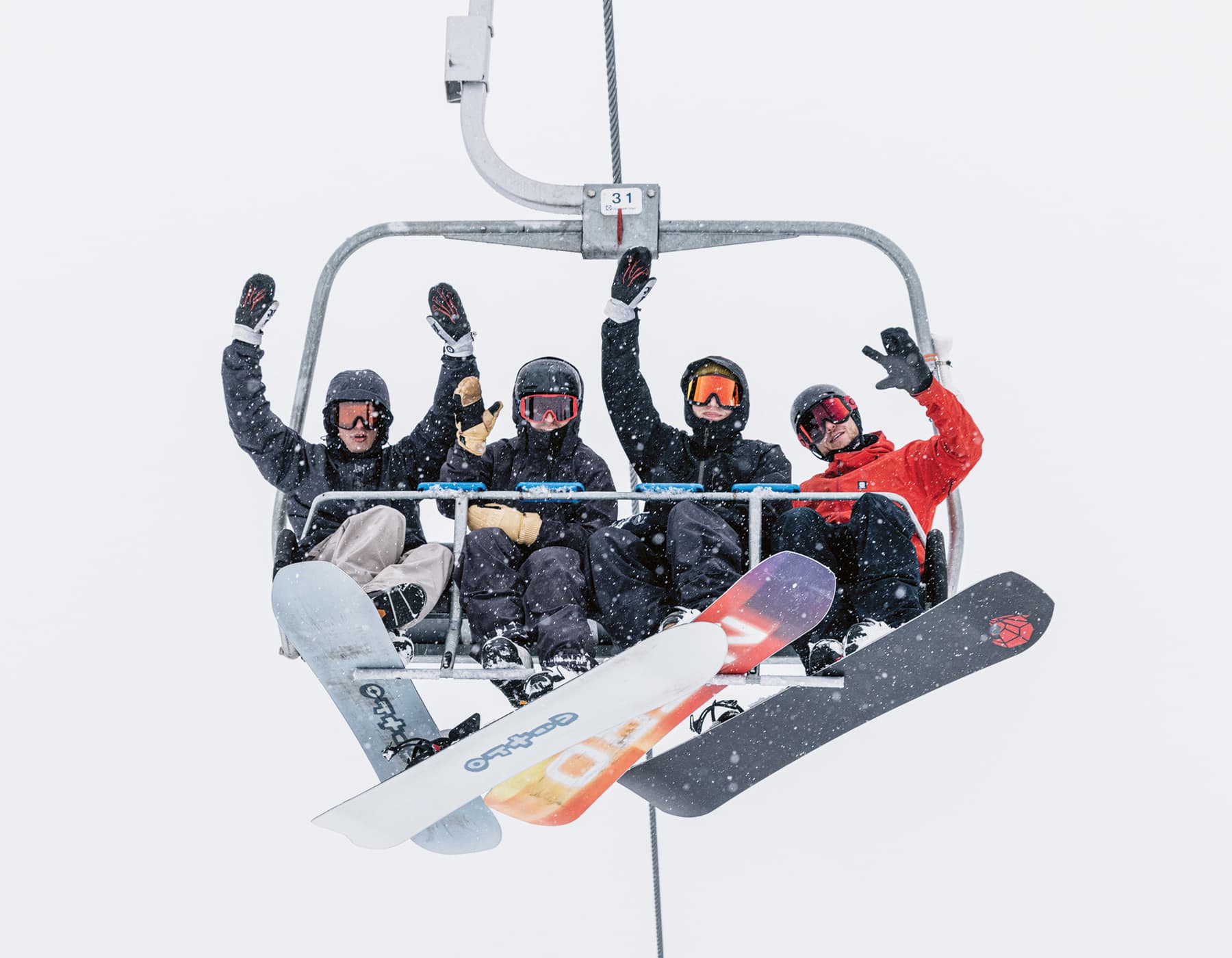 Four snowboarders sitting on a chairlift, with Nitro snowboards strapped to one foot. It is snowing and behind them is white sky. They are waving at the camera below them.