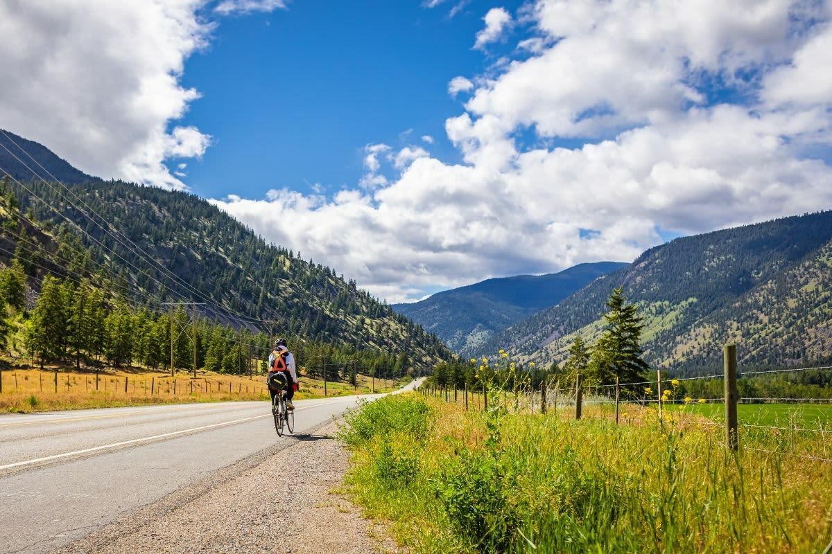 A cyclist heads down a quiet stretch of highway through the Okanagan backcountry, mountains rising on both sides and not another rider in sight.
