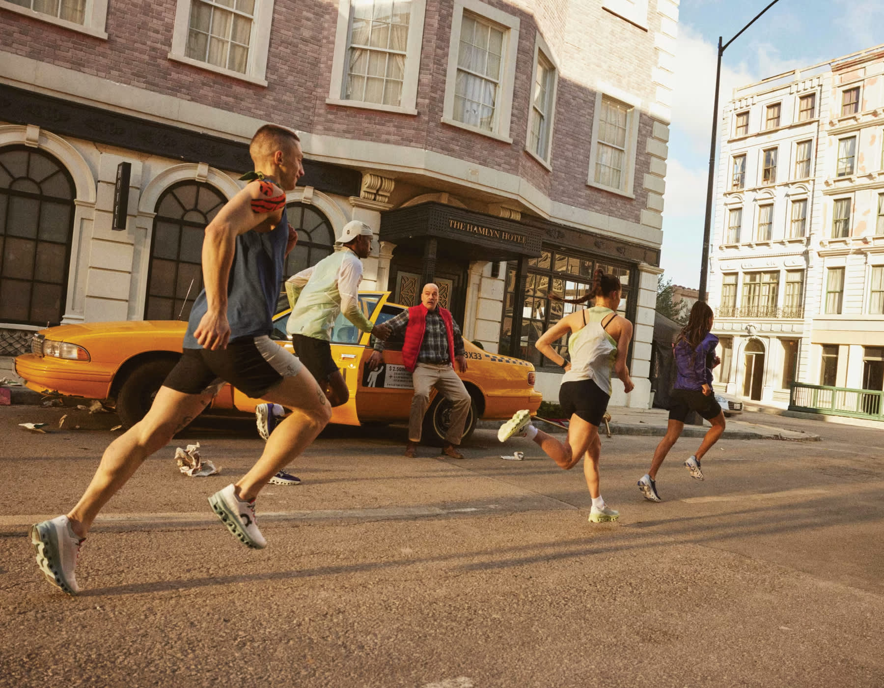 A group of people jogging down the road past brick buildings and a man exiting a yellow taxi car.