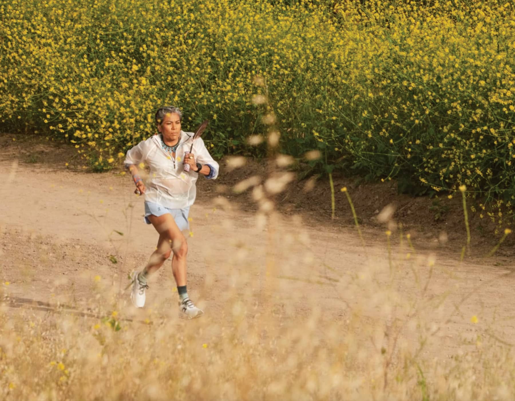 A Woman on a run wearing on shoes, running shoes and a windbreaker. Running on a dirt road with yellow flowers along the side. 
