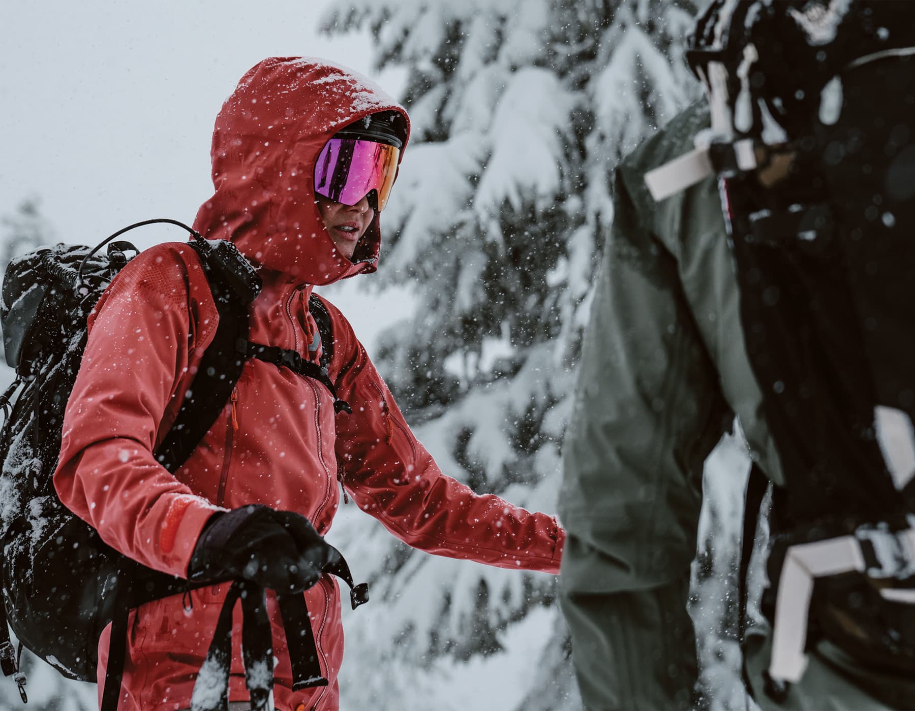 Two skiiers standing and talking to one another in heavy snowfall. One female with a red Orage jacket, backpack and goggles holding ski poles looking in the direction of another skiier with his back to the camera wearing a green Orage jacket and a pack pack