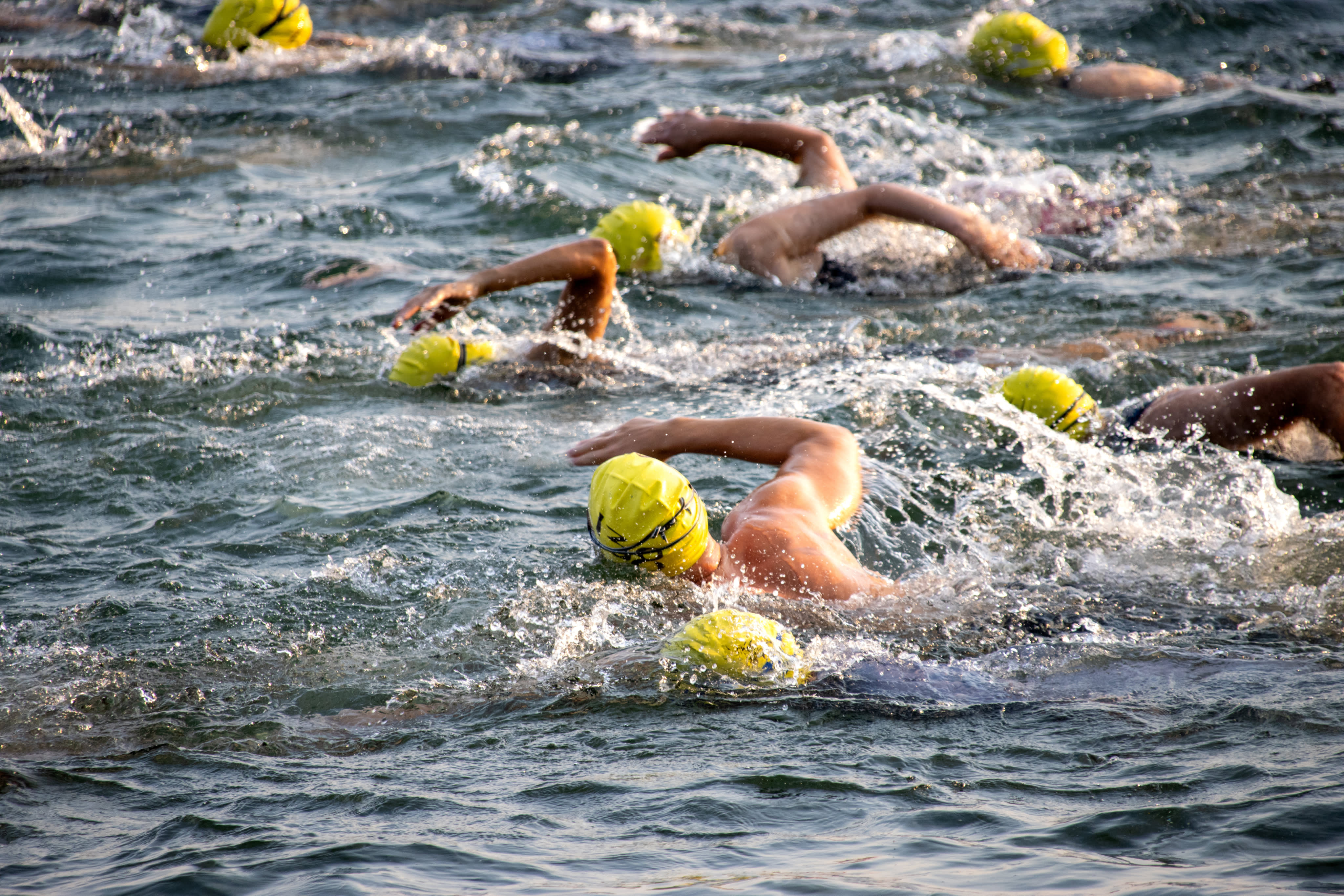 Open water swimmers in yellow caps mid-stroke, arms raised above choppy lake water during a triathlon swim leg.