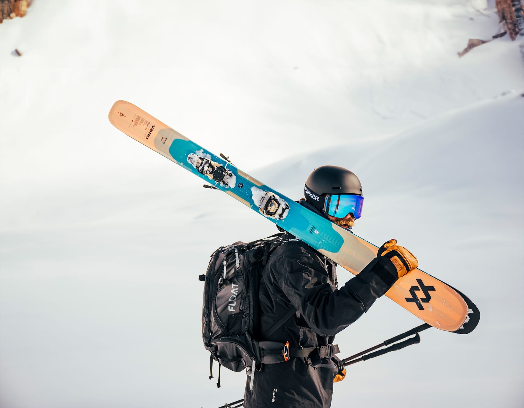 Skier holding a pair of Völkl skis on their shoulder, standing on fresh powder at the top of a snowy mountain