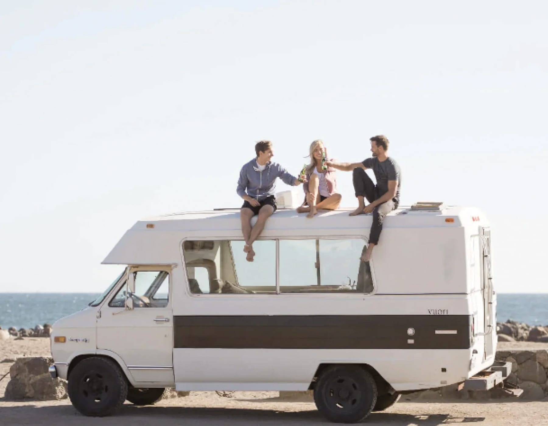 Three people sitting on top of a travel vac having a drink and doing cheers. The background is a beach with sand, water and blue sky.